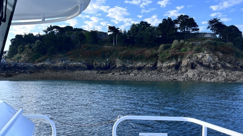 View from a boat of a rocky shoreline with trees and a blue sky.