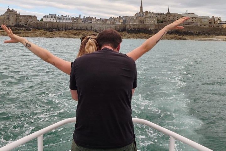 Couple on boat with arms wide open, facing cityscape across water.