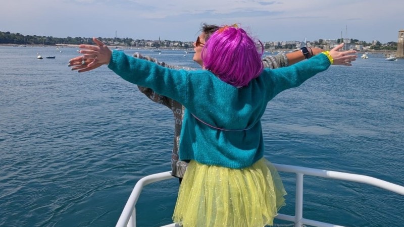 Person in a colorful wig and outfit spreading arms on a boat, overlooking the sea.