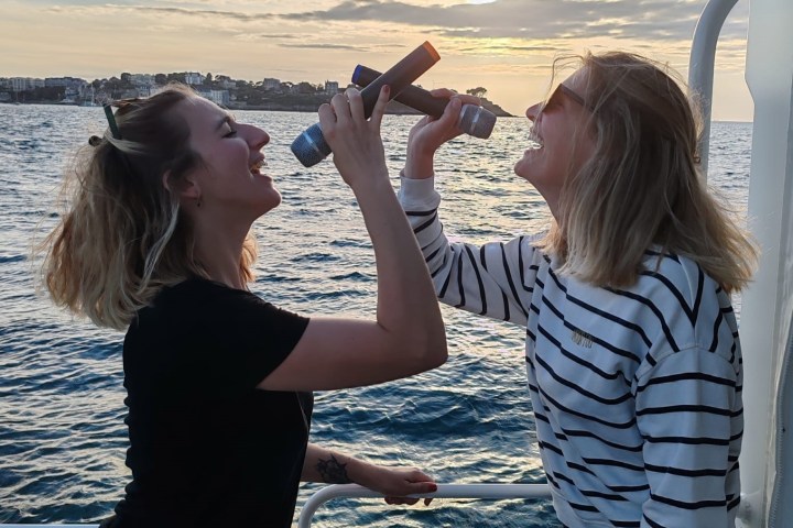 Two women laughing with metal objects by the sea at sunset.