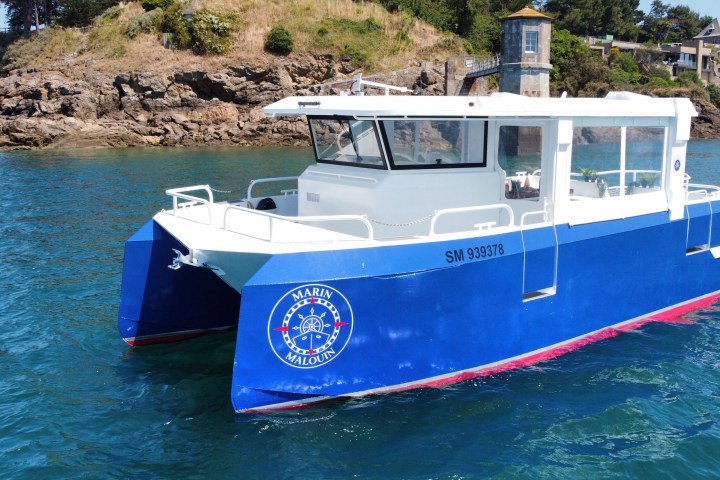 Blue catamaran in water near rocky shore and small building.