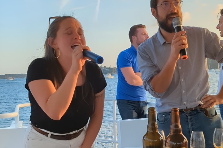 Two people singing into microphones on a boat with drinks on a table.