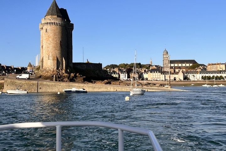 Stone tower and boats on water with town in background under clear blue sky.
