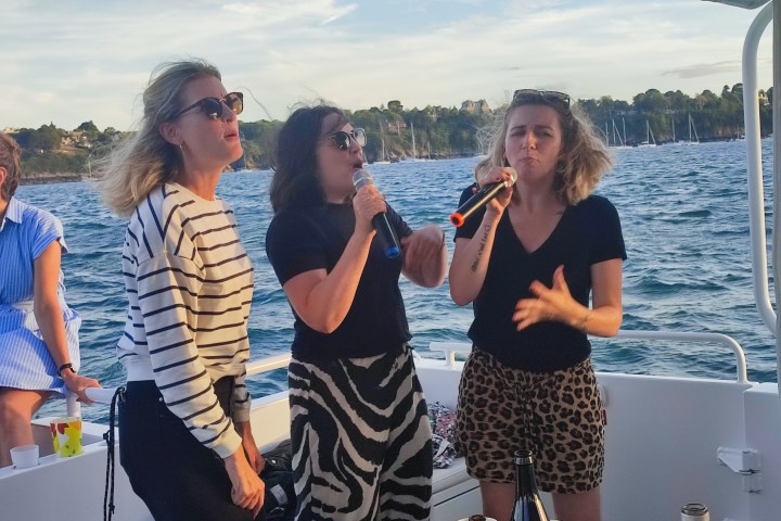 Three women singing on a boat with a scenic ocean backdrop.