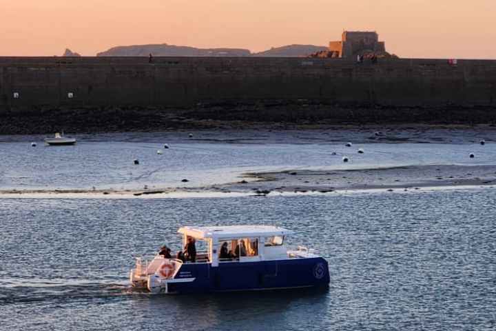 Small boat on calm water at sunset with distant shore and orange sky.