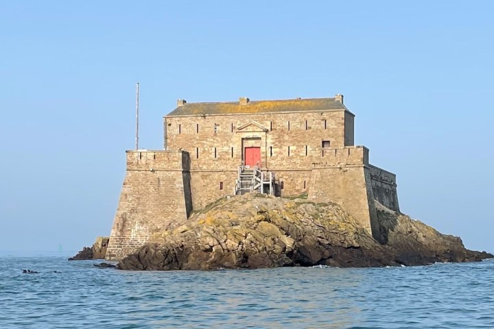 Stone fortress on rocky island surrounded by water under blue sky.
