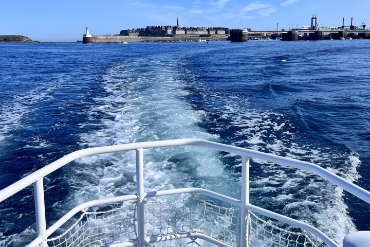 Boat railing and wake with cityscape and blue sky in background.