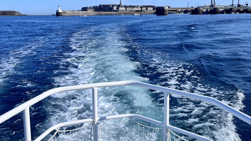 Boat railing and wake with cityscape and blue sky in background.
