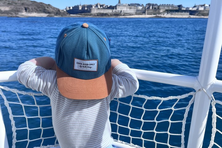 Child in a cap looking out at water from a boat, with a distant shoreline in view.