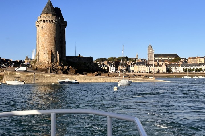 View from a boat of a coastal town with a round tower and sailboats.