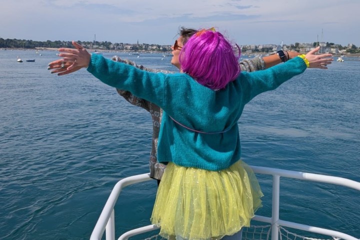 Two people enjoying a Titanic pose on a boat with blue sea and distant cityscape.