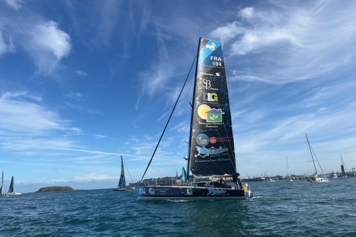 Sailboat with sponsor logos on a sunny sea, other boats in background.