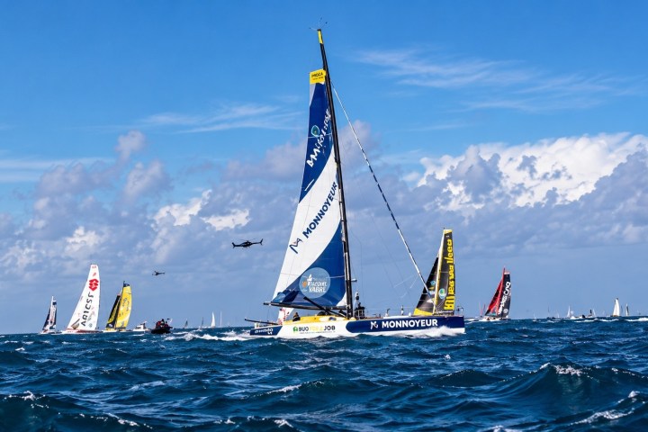 Sailboats racing in open sea under a cloudy sky, with a biplane flying overhead.